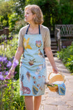 Model wearing the blue apron in the sunlit garden at Anne Hathaway's Cottage. She's holding a basket with a blue kitchen towel in it.