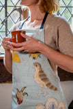 Model wearing the blue apron, holding a brown, glazed slipware cup. The hawk design motif is prominent.