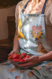 The model is wearing the blue apron, sitting on the steps in the kitchen of Anne Hathaway's Cottage. She's holding a brown slipware bowl of red strawberries in her hand. The sun is shining through the window emphasizing the embroidered flower pattern.