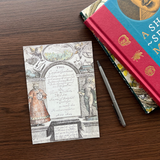 Postcard with old book title page on a dark brown table, placed next to a pen and books.