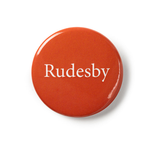 Red button badge with traditional pin fastening. White text says 'Rudesby'.