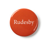 Red button badge with traditional pin fastening. White text says 'Rudesby'.